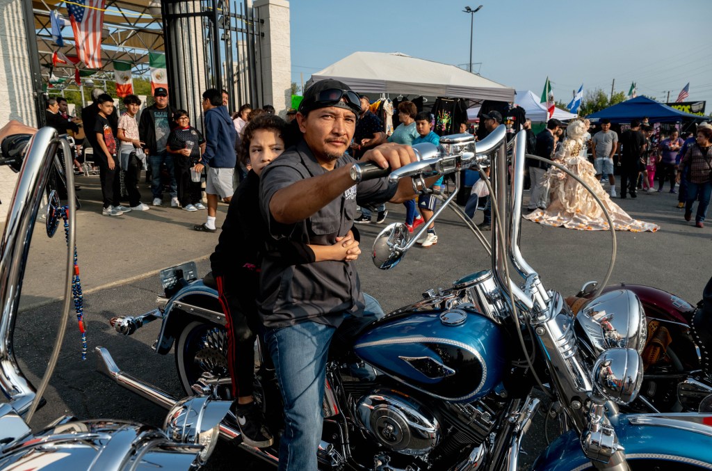 A man and child sit on a blue and chrome motorcycle with a crowd of people in the background.