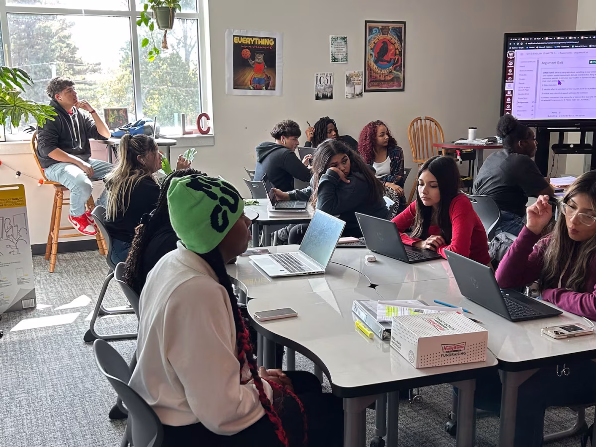High school students sit around tables in a sunny classroom, most with laptop computers in front of them.