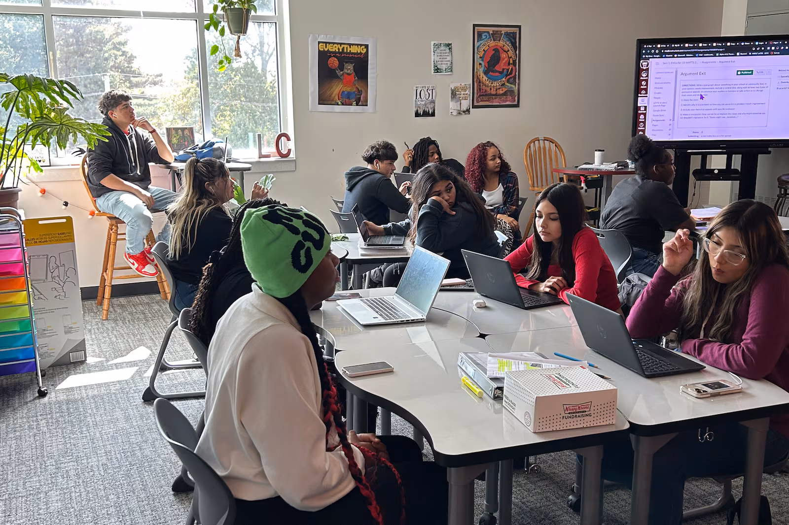 High school students sit around tables in a sunny classroom, most with laptop computers in front of them.