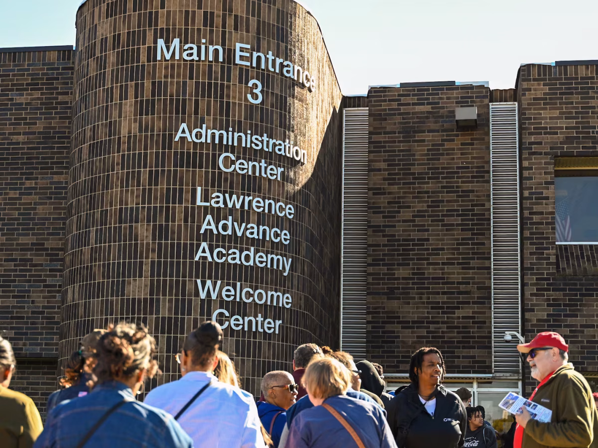 A crowd of people stand outside a brick building with white lettering that reads, "Main Entrance 3," "Administration Center," "Lawrence Advance Academy," "Welcome Center."