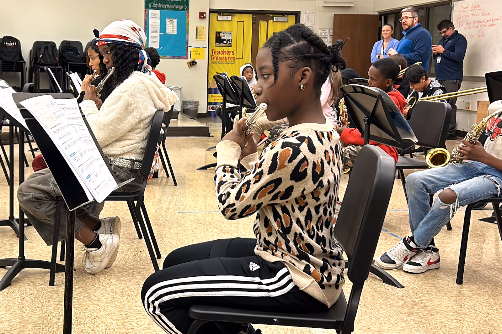 A seventh-grade girl sits in a band classroom with rows of other students, playing a flute and looking at music on a stand. A few more flute players are in the row with her, and some saxophones and brass players behind her.