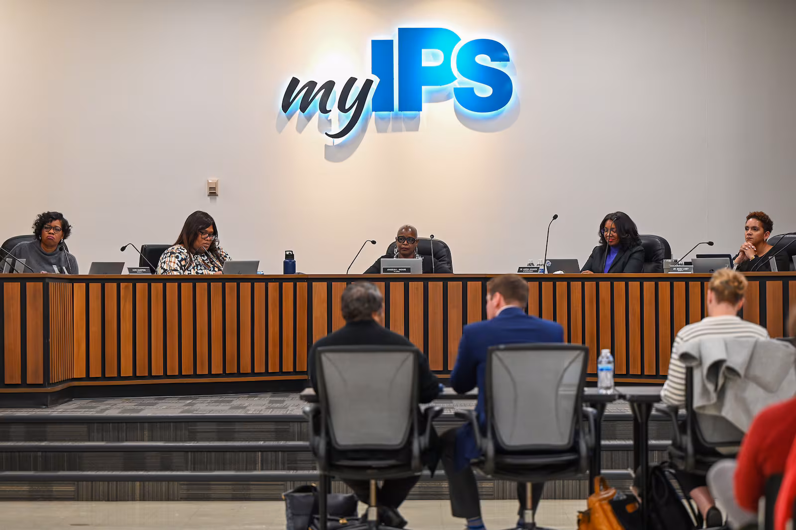 School board members sit at a curved desk on a platform under a lighted sign reading, "my IPS."