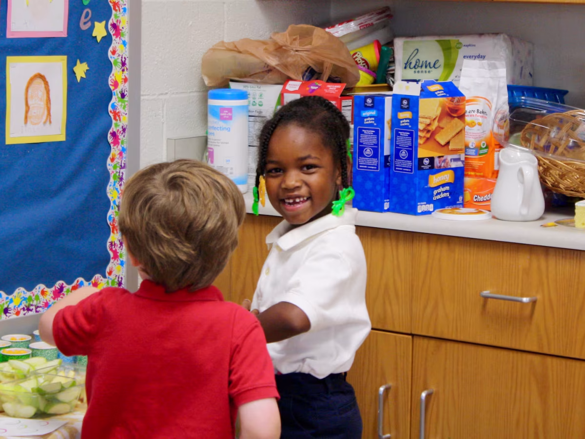 Two young children stand near a snack table in a classroom, one of them looking back at the camera and smiling.
