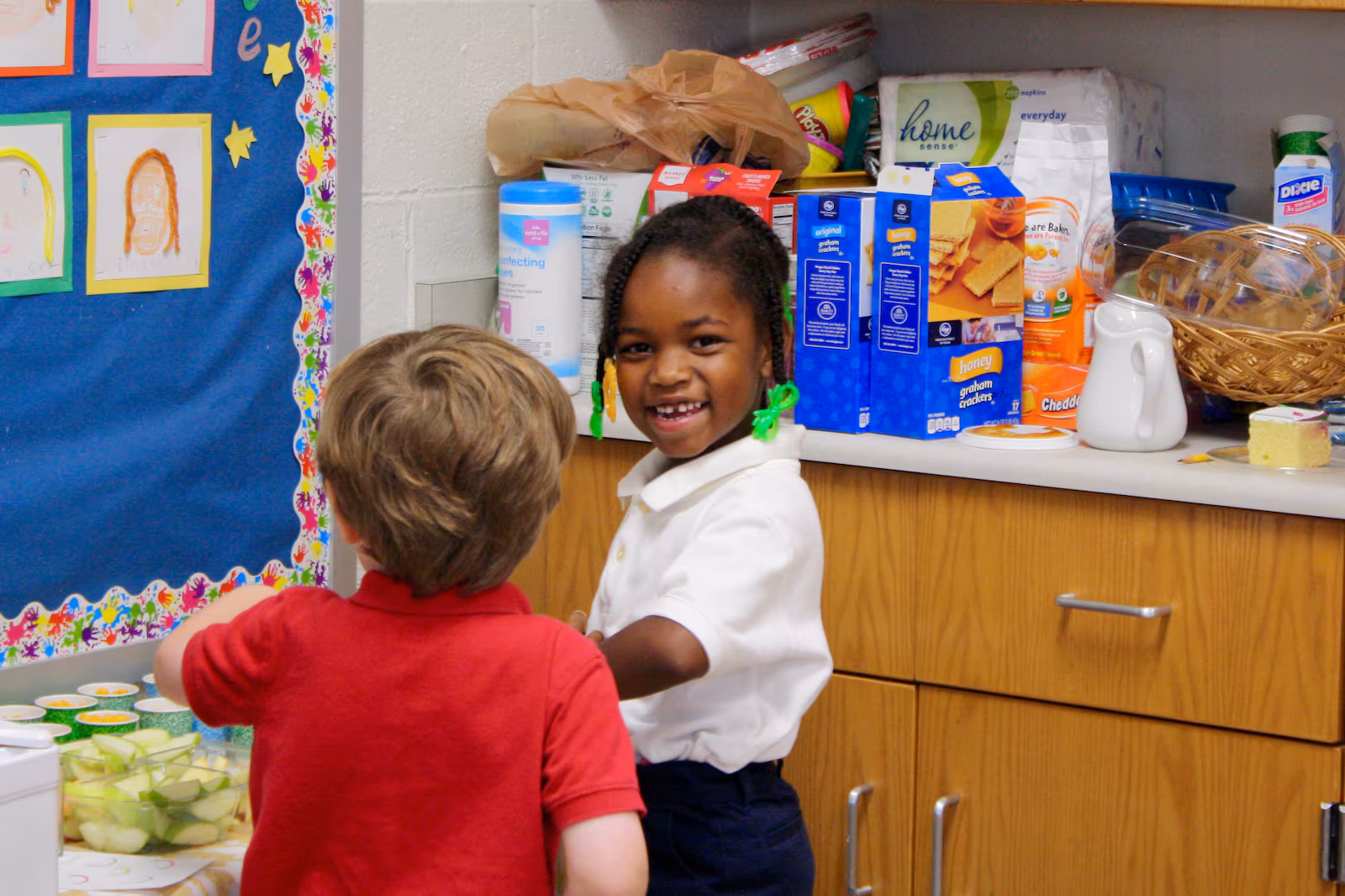 Two young children stand near a snack table in a classroom, one of them looking back at the camera and smiling.