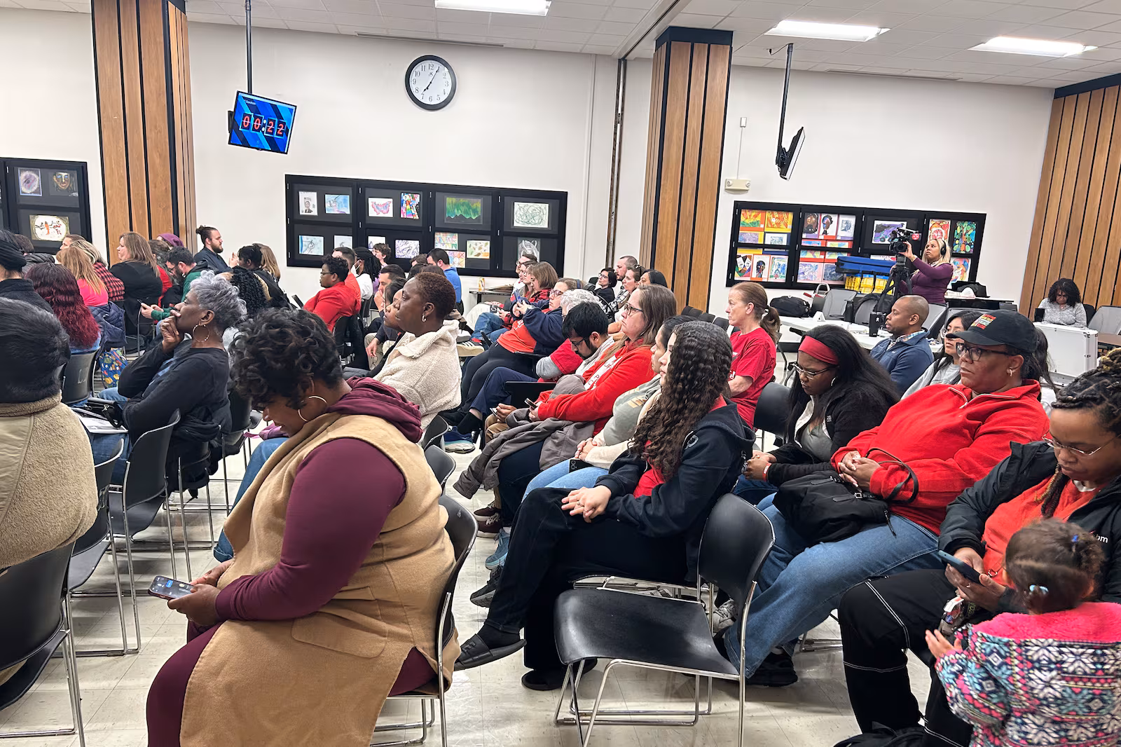 People fill rows of chairs in the audience of a school boar meeting, listening attentively.