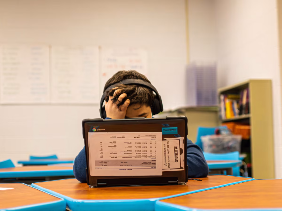 A young student sits at a laptop in a classroom, headphones on and head resting in one hand.