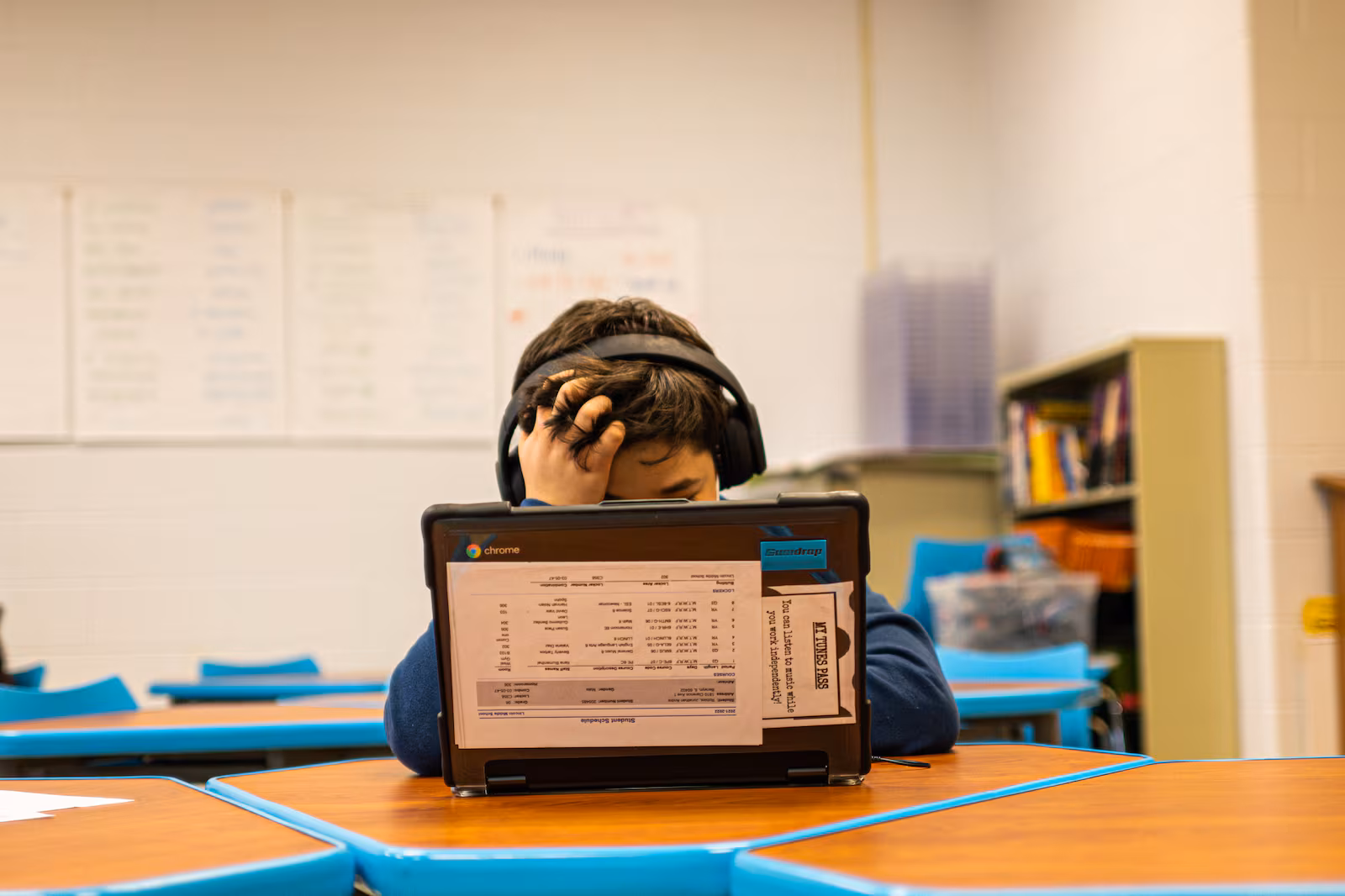A young student sits at a laptop in a classroom, headphones on and head resting in one hand.