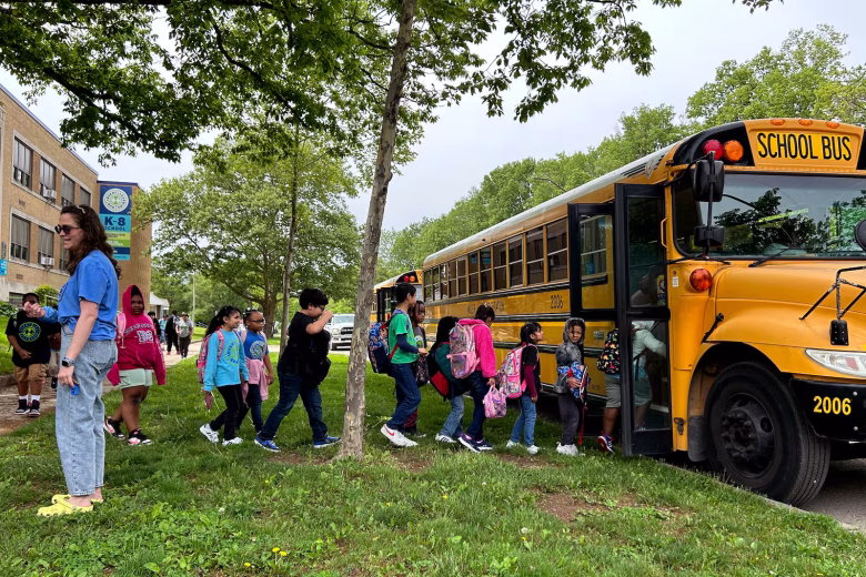Children line up to board a yellow school bus, while an adult looks on. They're in front of a brick school building.
