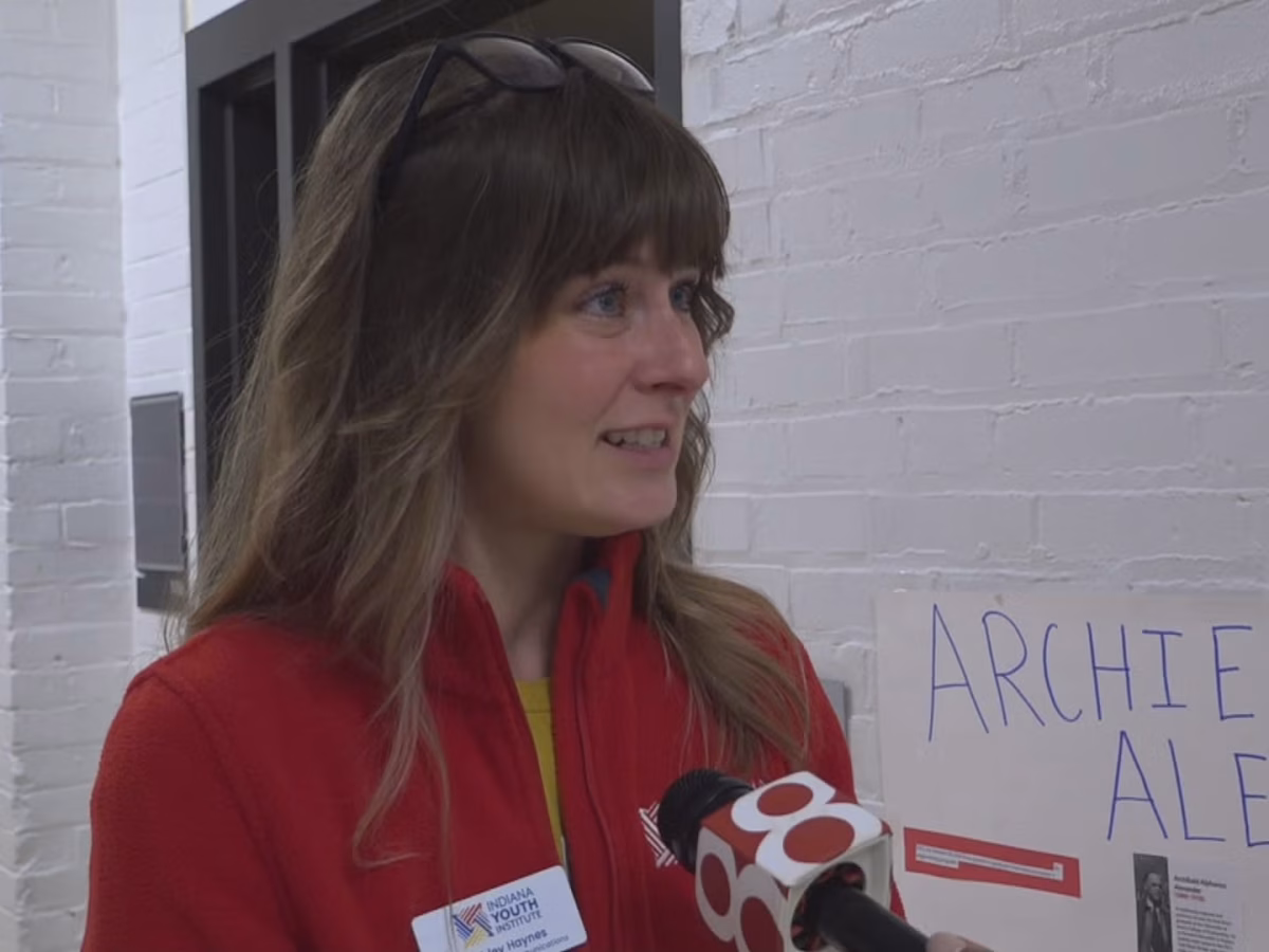 Ashley Haynes, who has light brown hair and a red fleece, speaks into a Channel 8 news microphone while standing in a school hallway.